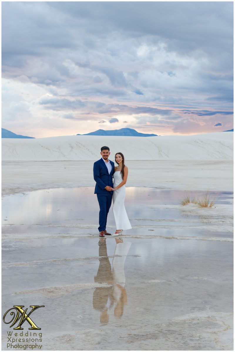 Engaged couple standing on white sand dunes under a vibrant sunset.