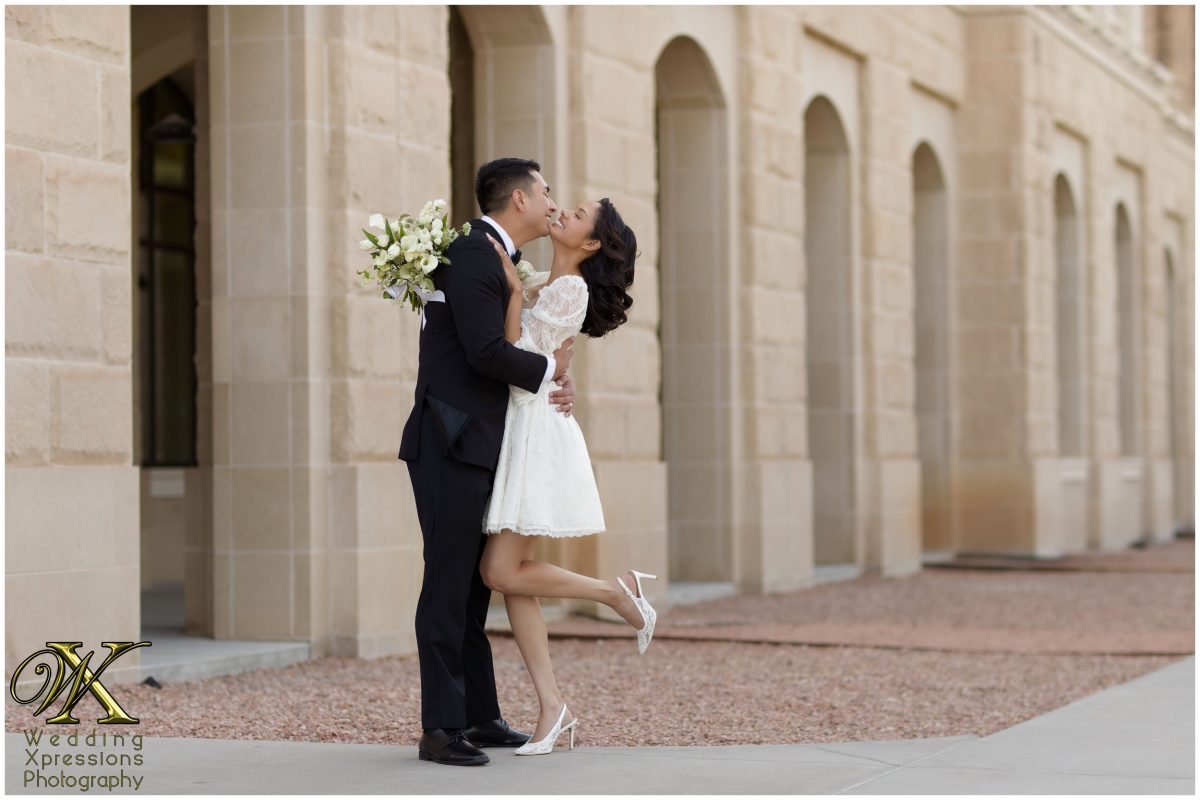 Bride wearing a chic above-the-knee white dress and groom in a black suit sharing a kiss in El Paso.